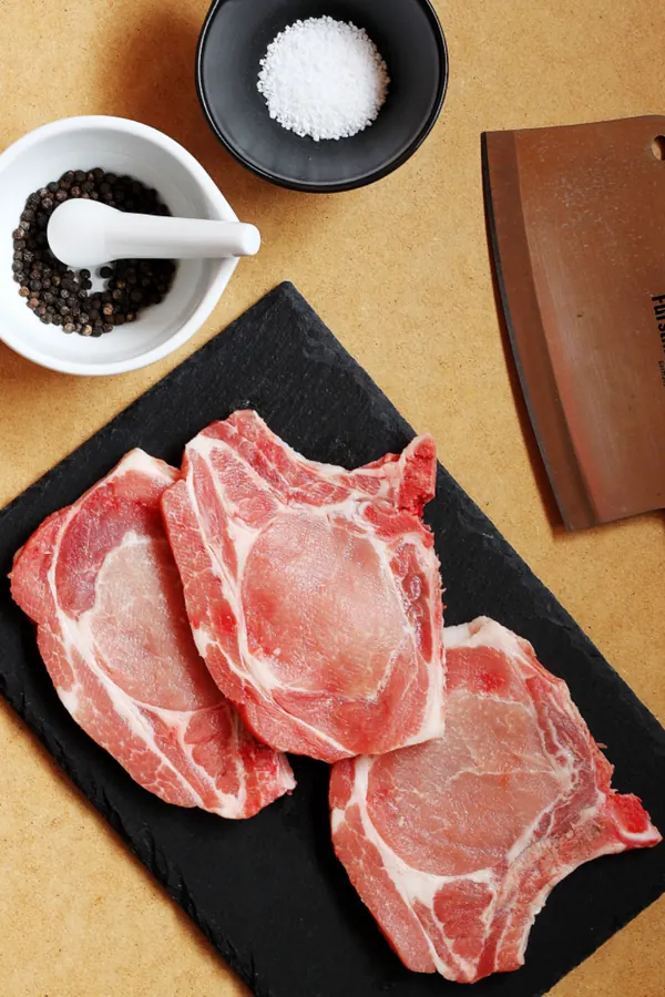 Raw bone-in pork chops on a black tray with salt and pepper